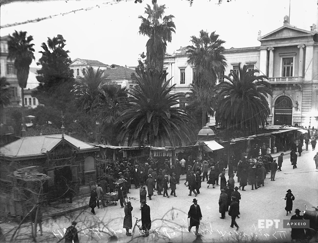 Άποψη της Πλατείας Δημαρχείου, σημερινή Πλατεία Εθνικής Αντίστασης, με υπαίθρια μαγαζιά. Στο βάθος το Μέγαρο της Εθνικής Τράπεζας επί της οδού Αιόλου. Δεκαετία 1920