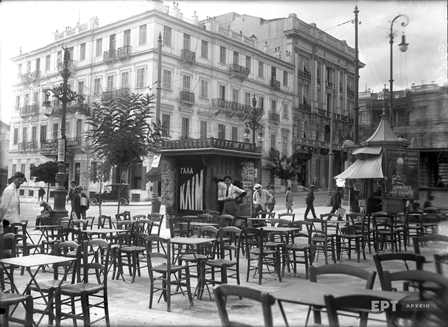 Άποψη της Πλατείας Συντάγματος προς την πλευρά της οδού Ερμού και του ξενοδοχείου Grand Hotel d’ Angleterre. Σε πρώτο πλάνο τα τραπεζάκια του καφέ-ζαχαροπλαστείου του Ζαβορίτη που στεγαζόταν στο ισόγειο του Μεγάρου Κορομηλά. Δεκαετία 1920
